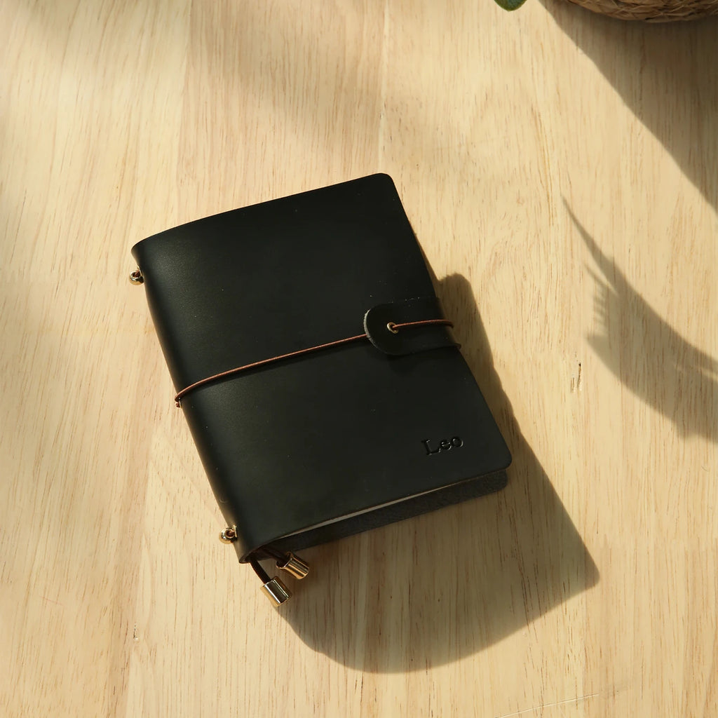 Midnight black passport holder with wrap strap and metal corner clasps, engraved logo
