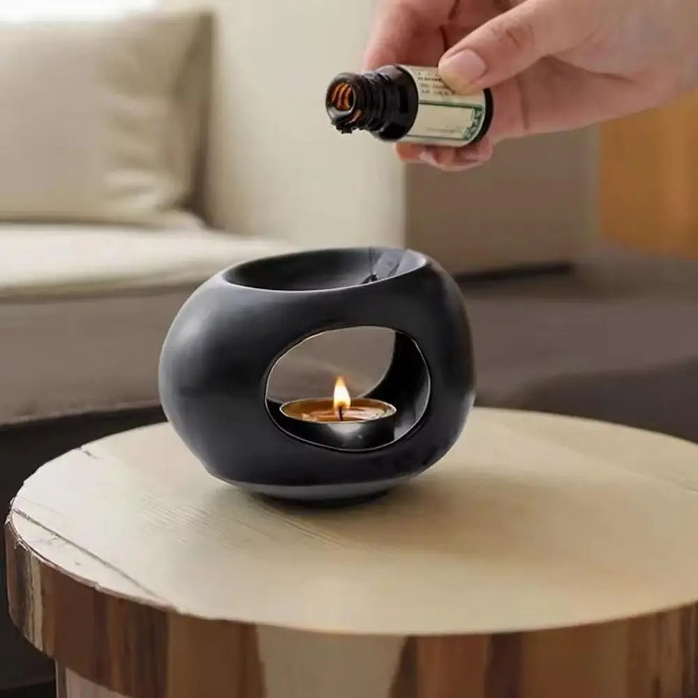 Black ceramic aroma oil burner on table with person pouring oil, flame visible