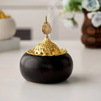 Ceramic oud burner with black dome base, gold openwork lid, visible incense smoke