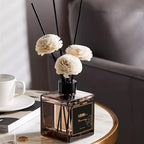 Square glass reed diffuser bottle with black reeds and white flower pom‑poms on table