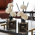 Two mini reed diffusers on tray: square bottles with black reeds and white pom‑pom flowers