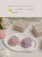 Two peony sculptural candles on a tray: light pink and lavender, with gift boxes nearby