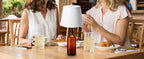 Wine bottle wireless LED lamp with white fabric shade on outdoor table, amber bottle