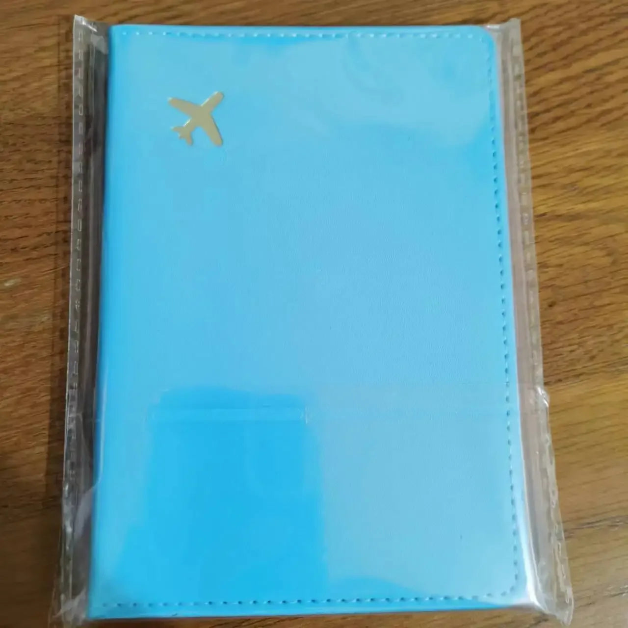 Sky blue passport sleeve in clear packaging, gold plane logo on front