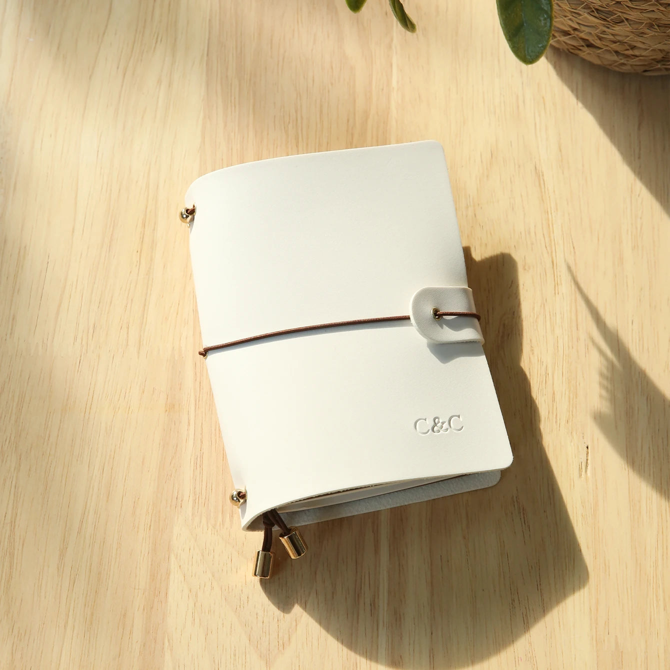 Ivory white passport holder with wrap strap and gold-tone corner clasps, engraved logo