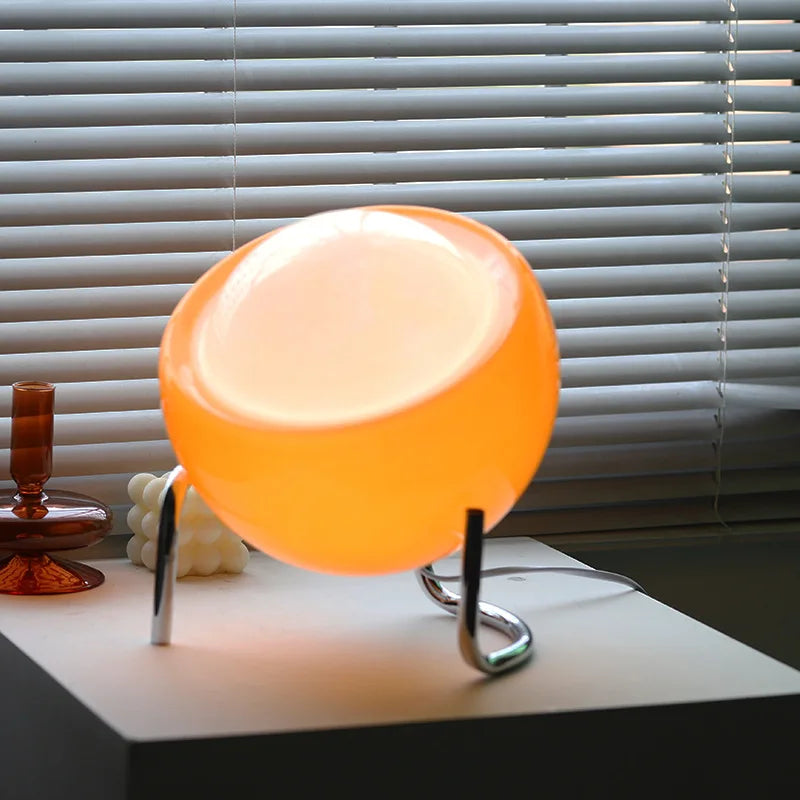 Glass globe table lamp with warm orange glow and chrome curved legs on tabletop