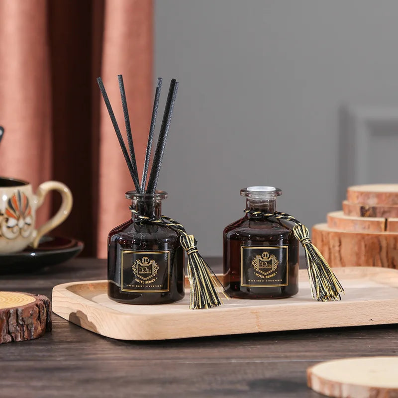 Two dark glass reed diffuser bottles with black reeds and gold tassels on light wood tray