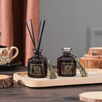 Two dark glass reed diffuser bottles with black reeds and gold tassels on light wood tray