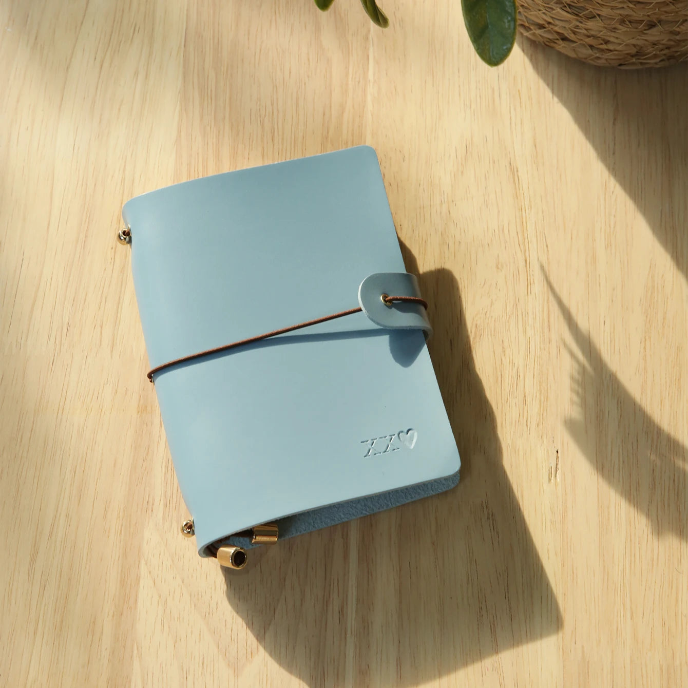 Light blue passport holder with wrap strap and metal corner clasps, engraved logo