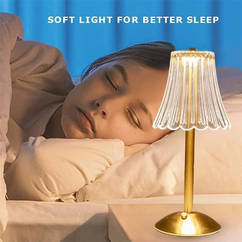 Cordless table lamp by bed with warm-lit pleated glass shade on gold base