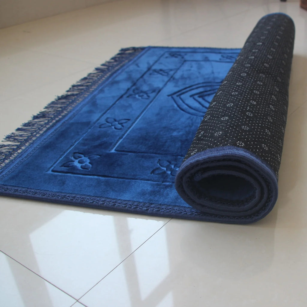 Velvet padded prayer rug in blue with raised arch pattern, rolled side visible, fringed border.