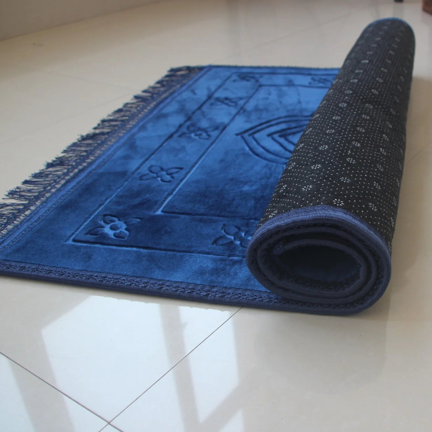 Velvet padded prayer rug in blue with raised arch pattern, rolled side visible, fringed border.