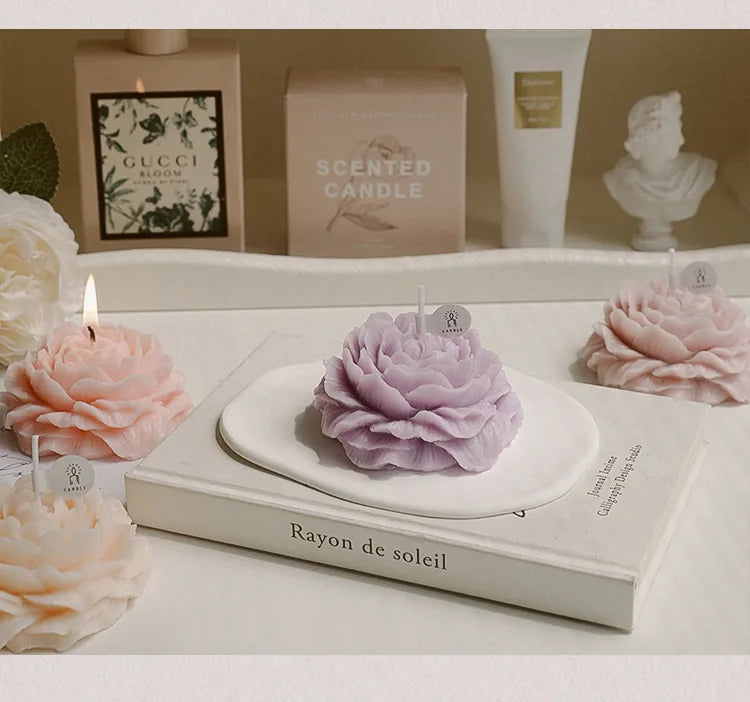 Purple sculptural peony candle with lit wick on a box, surrounded by peach and lilac