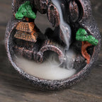 Close-up of resin waterfall incense burner: orange-roof house, green tree, flowing smoke waterfall